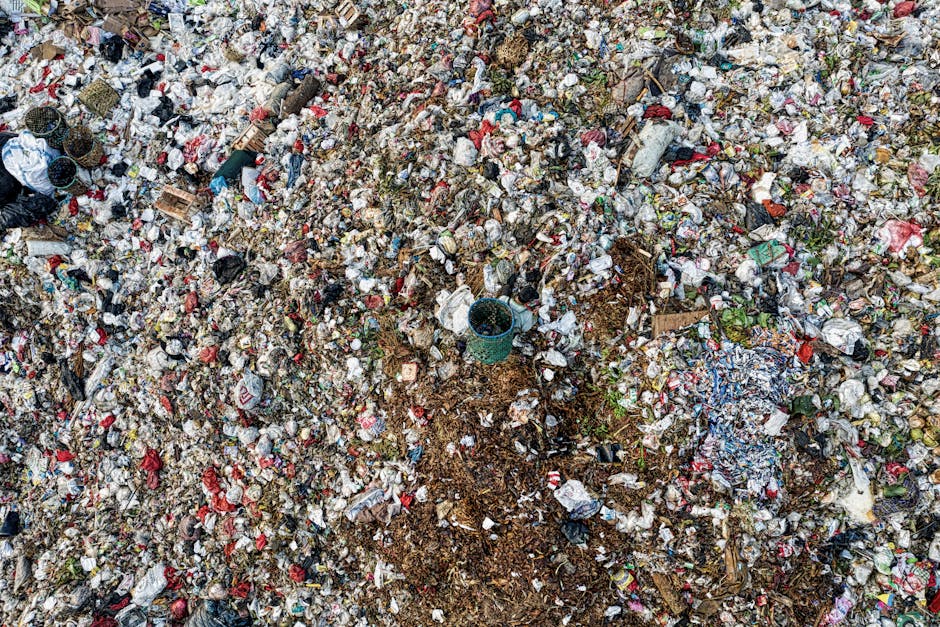A large, uneven expanse of mixed municipal and domestic waste spread across an outdoor area, showing a variety of materials including crumpled plastic bags, paper cartons, and miscellaneous packaging. The debris features different colors, predominantly reds, whites, and browns, with some visible green and blue plastics. There are small piles of soil and organic matter interspersed with the waste, and a black plastic bucket partially buried among the rubbish. The ground beneath the refuse appears muddy and uneven, with some scattered bits of vegetation. The scene suggests accumulated refuse from local waste collection or improper disposal in a private setting, with no clear waste segregation, typical of an area requiring rubbish removal services like those offered by Waste Disposal Finchley. The overall impression is of a cluttered, unmanaged waste site, possibly the result of a bulky waste removal or on-site clearance process, illustrating the importance of professional rubbish disposal for environmental management.