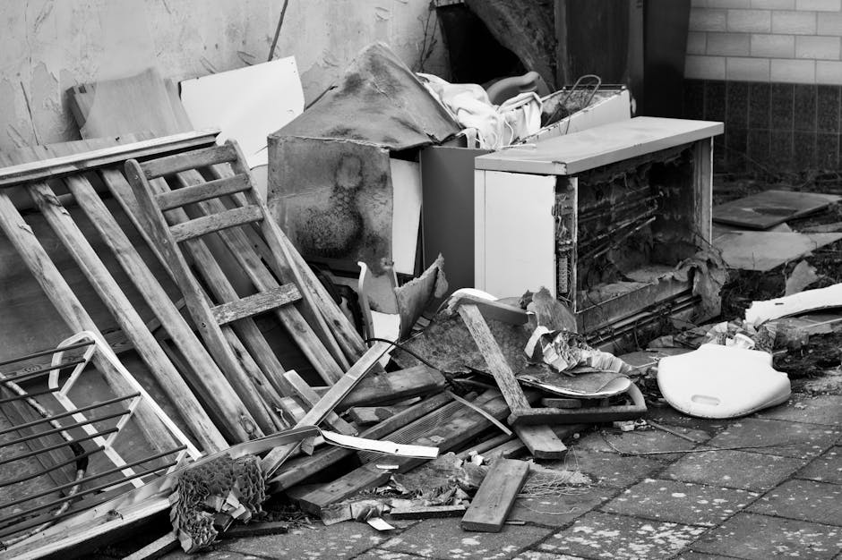 A cluttered outdoor scene featuring discarded wooden furniture, including a collapsed wooden chair or bed frame with slatted support, resting on a paved surface. Adjacent to the furniture is a damaged and partially disassembled white cabinet or appliance, with visible dirt and rust stains, leaning against a wall. Surrounding the main objects are various debris, including broken wood pieces, a piece of textured fabric, and scattered small fragments of construction or household waste. To the right, there is a white plastic container or seat, and the background reveals a mix of brick and concrete wall surfaces. The area appears to be an unmanaged or neglected space, indicative of a spot where private waste or bulky items are temporarily stored for removal, aligning with services provided by Waste Disposal Finchley for rubbish and waste clearance in residential or commercial settings.