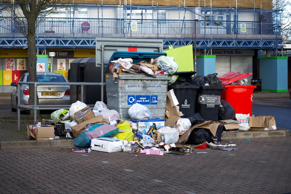 A large collection of mixed household and office waste, including cardboard boxes, plastic bags, paper, and packaging materials, piled up around a metal bin with a partially open lid, situated on a paved area in an urban environment. The waste extends onto the surrounding pavement and partially blocks a pathway, with various items spilling onto the ground. Behind the pile, a silver hatchback car is parked adjacent to a metal railing. In the background, there is a commercial building with storefronts, featuring signs and awnings, and a section of blue scaffolding or construction equipment indicating ongoing maintenance or renovations. The scene is illuminated by natural daylight, highlighting the disorganized state of the rubbish, which is representative of an unmanaged or excessive waste collection situation that could be addressed through private rubbish removal services like those offered by Waste Disposal Finchley. The overall setting emphasizes the importance of proper waste disposal in residential or commercial contexts, aligning with the scope of independent rubbish clearance options.