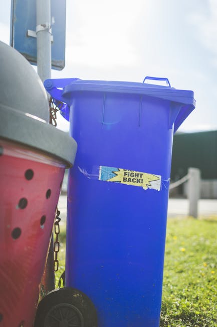 A tall, bright blue plastic rubbish bin with a closed lid and a small rectangular sticker that reads 'FIGHT BACK!' in yellow and black text, positioned outdoors on a grassy area. To the left of the blue bin is a partially visible red and black polka-dotted waste container with a grey lid, which appears to be made of plastic or metal. Both containers are placed on a paved surface, and in the background, there is a metal pole supporting a sign or structure, with a partly cloudy sky overhead. The scene suggests a residential or public space where waste is collected, highlighting options for private or alternative rubbish disposal services such as waste management companies like Waste Disposal Finchley, especially relevant to addressing collection delays or independent waste handling.