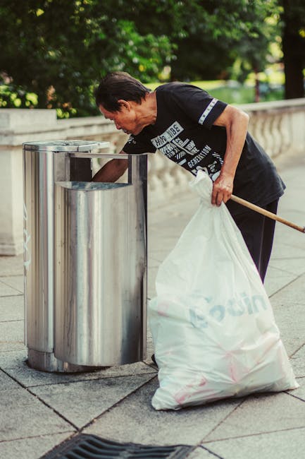A woman with short dark hair, wearing a black T-shirt with white and blue printed text, is disposing of waste into a modern, cylindrical metal litter bin situated on a concrete sidewalk. She is holding a large white garbage bag in her left hand, which appears to be filled with waste, and using her right hand to place rubbish into the bin. The bin has a sleek, stainless steel finish with a rounded opening and is positioned against a background of greenery and trees, suggesting an outdoor urban or park setting. The scene is well-lit, with natural daylight illuminating the woman, the metallic surface of the bin, and the surrounding environment. This image visually relates to private waste management and rubbish collection services, such as those offered by Waste Disposal Finchley, highlighting alternative waste handling practices in public outdoor spaces, including litter removal and on-site waste disposal efforts. The overall setting emphasizes cleanliness and proper rubbish separation within a community context.