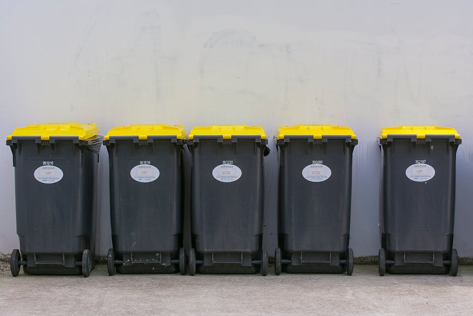 A tall, bright blue plastic rubbish bin with a closed lid and a small rectangular sticker that reads 'FIGHT BACK!' in yellow and black text, positioned outdoors on a grassy area. To the left of the blue bin is a partially visible red and black polka-dotted waste container with a grey lid, which appears to be made of plastic or metal. Both containers are placed on a paved surface, and in the background, there is a metal pole supporting a sign or structure, with a partly cloudy sky overhead. The scene suggests a residential or public space where waste is collected, highlighting options for private or alternative rubbish disposal services such as waste management companies like Waste Disposal Finchley, especially relevant to addressing collection delays or independent waste handling.