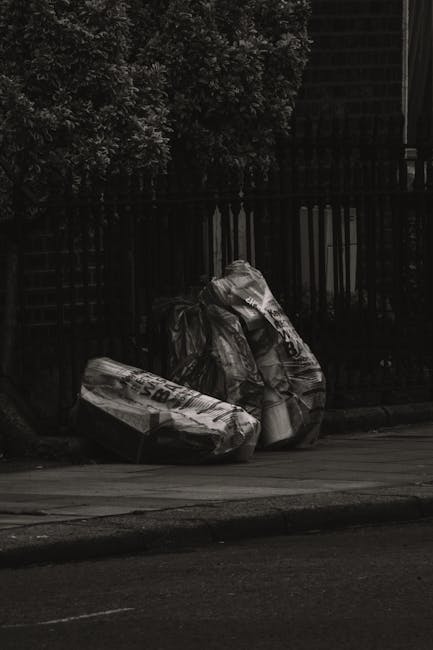 Three large black rubbish bags, made of thick plastic with visible creases and folds, are placed on the pavement next to a metal fence and a small tree with dense foliage. The bags appear to be filled with household or garden waste, and their surfaces reflect minimal light, emphasizing their crumpled texture. The setting is outdoors, possibly on a residential street, with a paved roadway in the foreground and a dark, vertical slatted fence behind the bags. The environment suggests a typical scene of voluntary rubbish disposal or collection, which could be associated with private waste removal services such as those offered by Waste Disposal Finchley. The subdued lighting and shadows create an overall neutral, utilitarian atmosphere, highlighting the disposal of waste materials awaiting collection or removal in a local urban area.