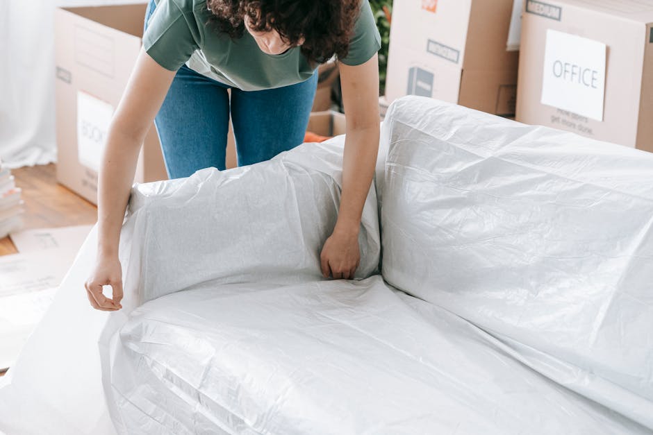 A person with curly dark hair, wearing a green t-shirt and blue jeans, is seen in the process of wrapping a large piece of furniture with a white protective cover made of plastic or fabric. The individual is standing on a wooden floor surrounded by cardboard boxes labeled with various categories, such as 'OFFICE' and 'MEDIA,' indicating a move or clearance activity. In the background, there are stacked boxes and furniture, suggesting an interior space being prepared for disposal or removal. The environment appears well-lit with natural or artificial light, highlighting the textures of the covering material and the surrounding items. This scene exemplifies a typical step in a professional rubbish removal or furniture clearance service, like those provided by Waste Disposal Finchley, aimed at safely wrapping and preparing bulky items for private disposal or alternative waste handling outside of local authority collection systems.