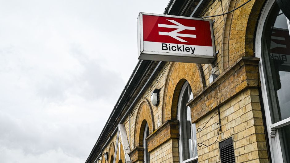 A close-up view of the exterior of a brick building featuring a prominent rectangular sign with a red and white logo of a railway, indicating Bickley station, mounted above a large arched window with white frames. The brickwork is a warm yellow-brown tone with visible mortar joints, and there is a small black vent alongside the window. The building's architectural details include rounded arches and decorative brickwork, suggesting an older construction style. Overhead, the sky appears cloudy with grey and white clouds, casting soft, diffused light over the scene. In the background, part of the building's roof and the edge of the station signage are visible. This setting relates to public transport infrastructure, and the image subtly aligns with the context of executing on-site or alternative waste disposal options, as Waste Disposal Finchley offers, by highlighting the external environment where rubbish removal services might be required near transport stations and surrounding areas.