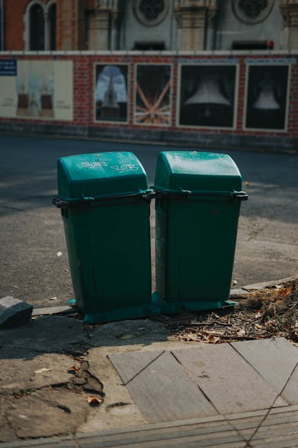 Two green plastic rubbish bins with hinged lids are positioned side by side on the edge of a sidewalk, with a small area of dried leaves and dirt around their bases. The bins have textured surfaces and are marked with white labels that include the words 'Rubbish' and a logo, indicating a private waste collection service. Behind the bins, there is a paved street with a slight curve, and further in the background, a brick building with large decorative windows and ornate architectural features can be seen. The scene is illuminated by natural daylight, casting soft shadows on the ground. This setup suggests a typical site for local refuse collection, potentially as an alternative to municipal waste disposal, reflecting the practice of private rubbish removal services like Waste Disposal Finchley. The environment appears urban, with the bins placed conveniently at the edge of the pavement for easy access and collection, exemplifying an organized approach to on-site waste management.